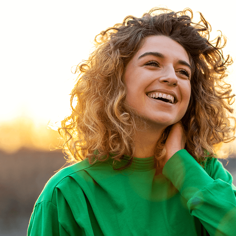 Smiling woman with short curly hair, wearing a green top, hand on neck, against sunset backdrop.