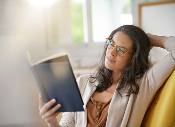 A woman with glasses reading a book on a yellow sofa, smiling with a sparkle in her eyes.