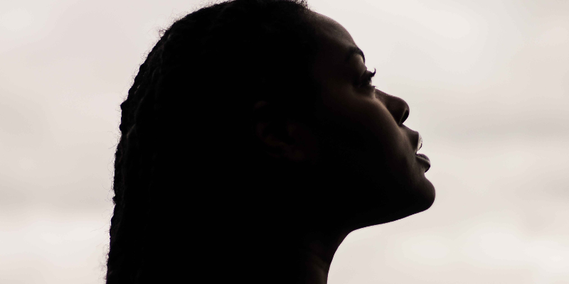 A dark, side-profile shot of a woman's head, looking upward with a slightly open mouth, against a blurry sky background.