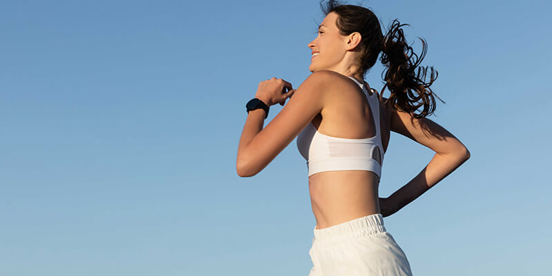 A woman jogging with a pleasant smile in a white sports kit, captured from a low side angle against a backdrop of clear blue sky.