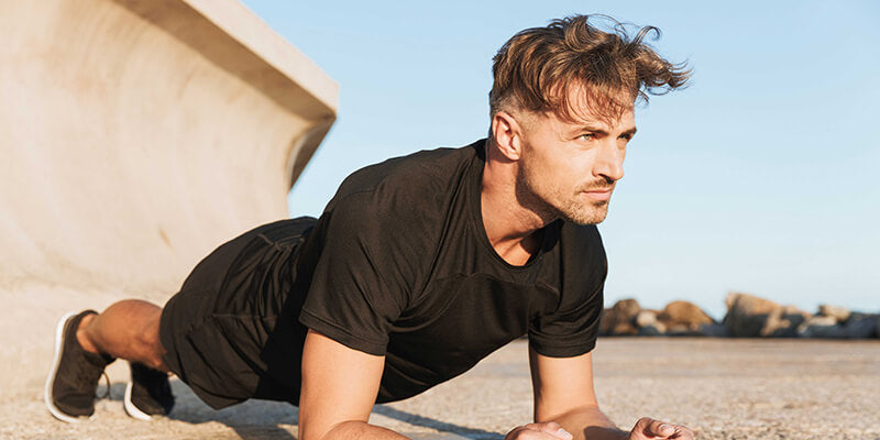 A man performing plank exercises on the sand in an outdoor setting during the daytime with sunshine.