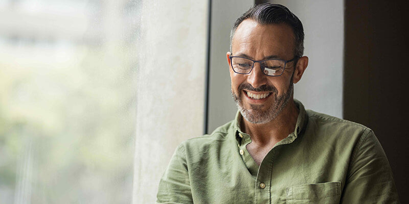 A medium close-up shot of a bearded man in a green shirt and glasses, smiling and looking downwards, possibly at a tablet.