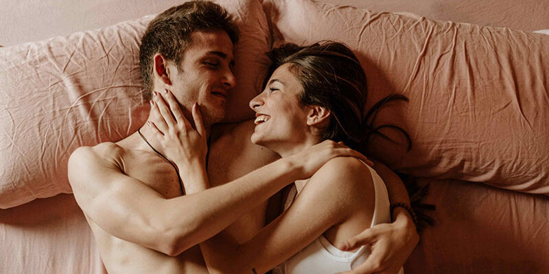 A man and woman hugging each other and lying on a bed, looking at each other's faces with smiles.