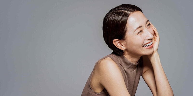 A medium close-up shot of a short-haired woman touching her face on the opposite side of the camera, smiling broadly.